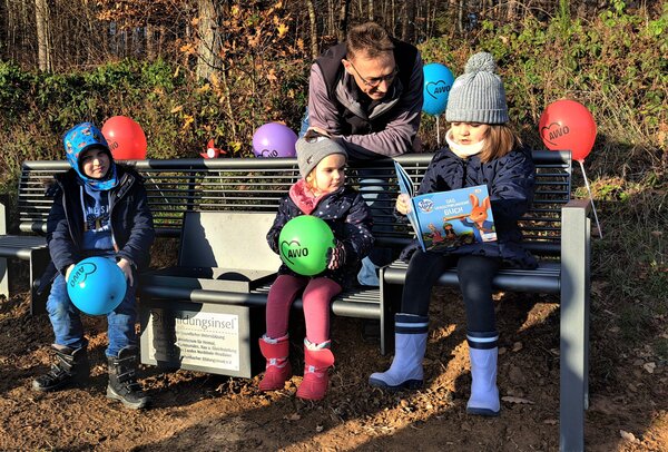Die Kindergartenkinder des AWO-Familienzentrums nehmen sofort Platz auf der neuen Bildungsinsel in Hadem. Auf dem Bild steht Tomas Irle hinter der Bank, auf der drei Kinder sitzen. Ein M&auml;dchen liest ein Buch. 