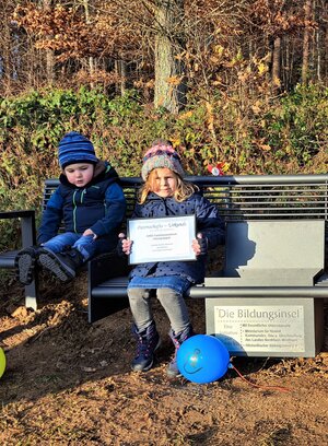 Bild vergr&ouml;&szlig;ern: Die Kinder des Hilchenbacher AWO-Familienzentrums &uuml;bernehmen die Patenschaft f&uuml;r die Bildungsinsel in Hadem. Daf&uuml;r haben sie eine Urkunde erhalten, die ein M&auml;chen in die Kamera zeigt. Sie sitzt neben einem Jungen auf der neuen Lesebank.