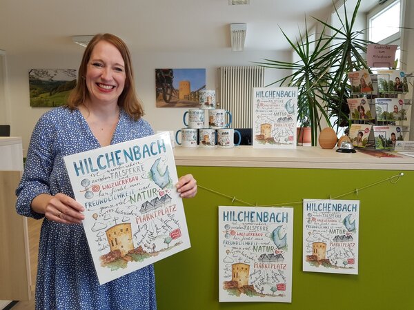 Lydia Huckenbeck zeigt das Hilchenbach-Poster, das in der Touristikinformation am Marktplatz erh&auml;ltlich ist. Auch Tassen mit dem von ihr gestalteten Motiv k&ouml;nnen hier erworben werden.